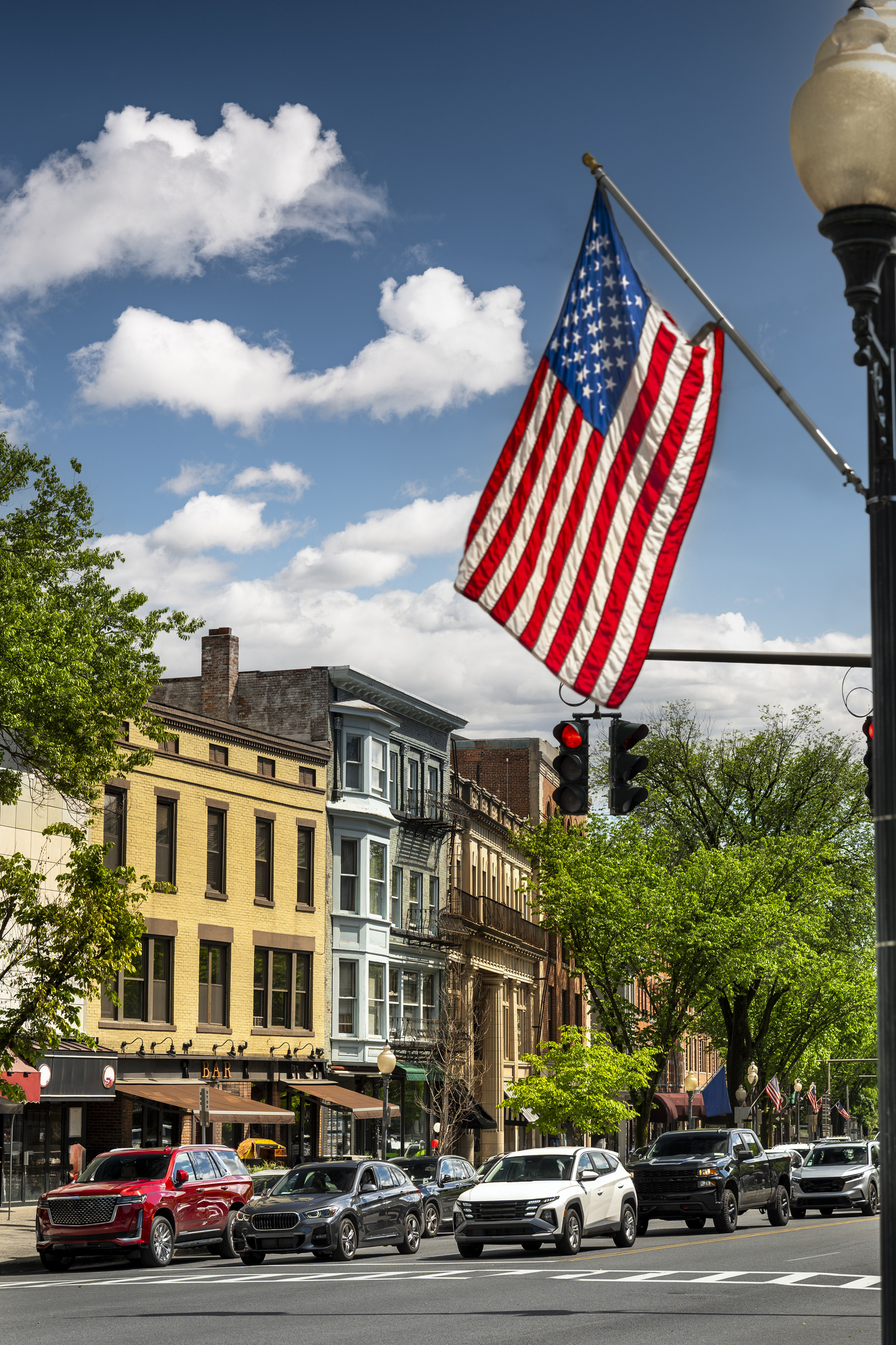 American flag flying proudly over an urban roadway with cars driving past historic multistory brick buildings on a bright summer day